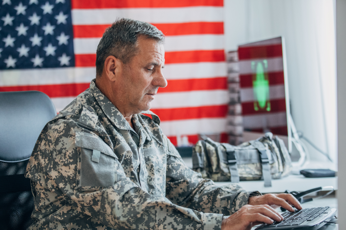 Older US soldier sitting at a desk on a laptop in front of a US flag
